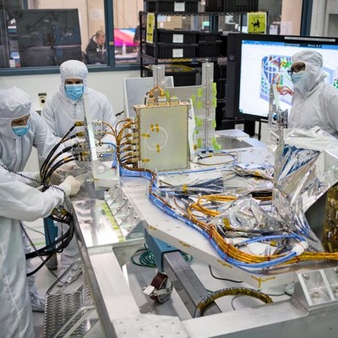 Engineers in full cleanroom garments working around instrumented aerospace hardware on a bench, with a wall display showing sensor data.