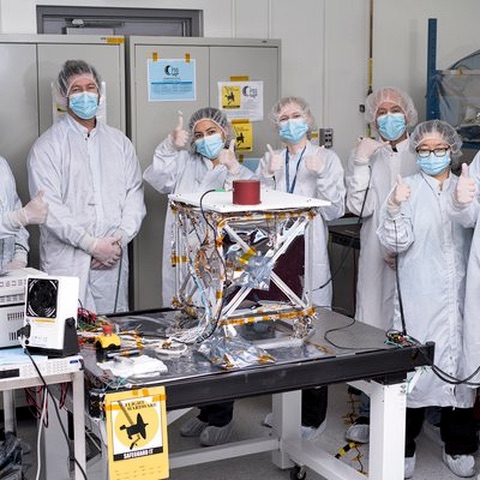 An engineering group in laboratory coats and hairnets standing around a foil-wrapped truss assembly on an anti-static bench.